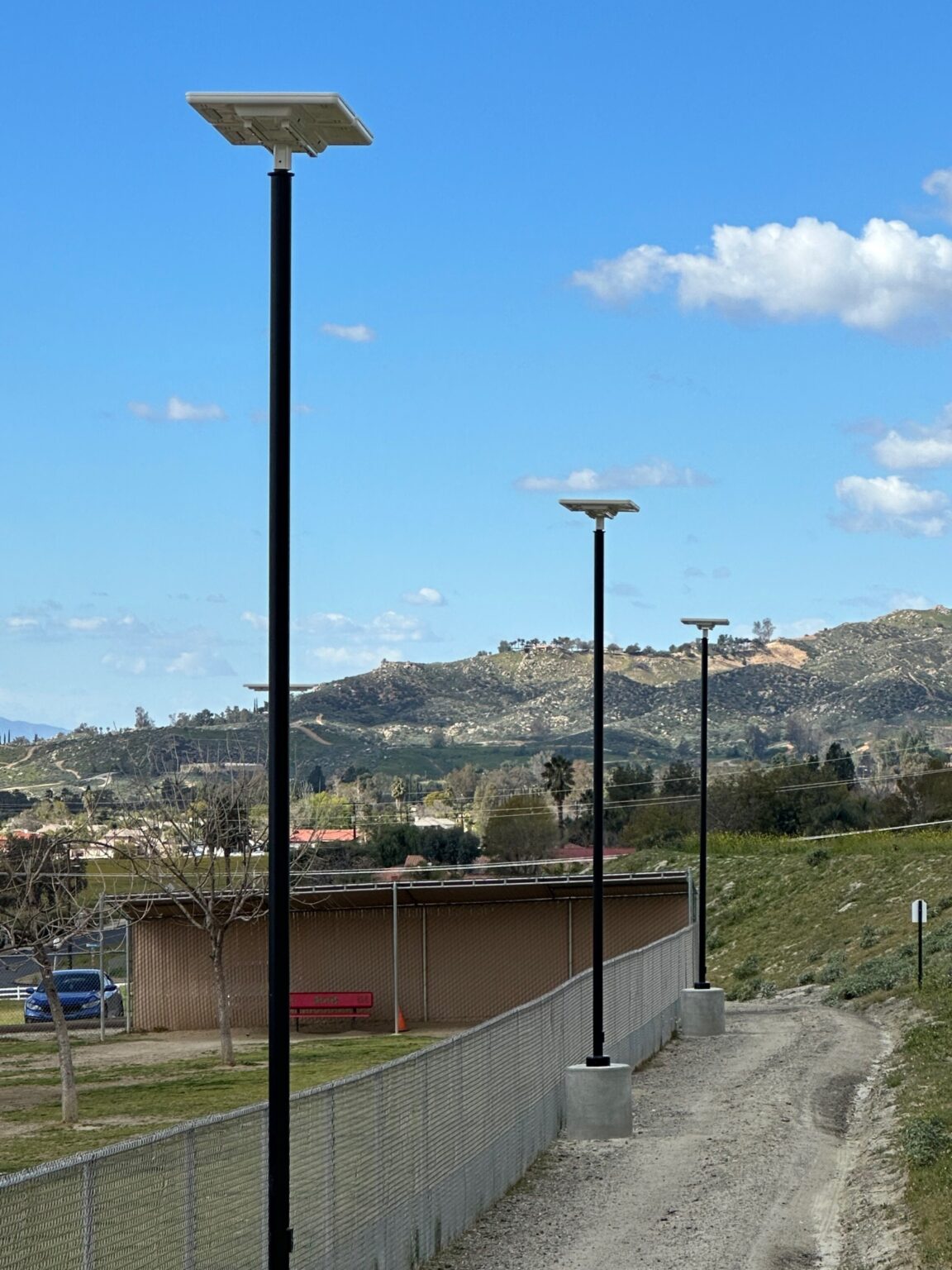 Three of Sol's iSSL solar lights lining a fence at a dog park in California with a covered shade area, parked car, and hills against a clear sky in the background