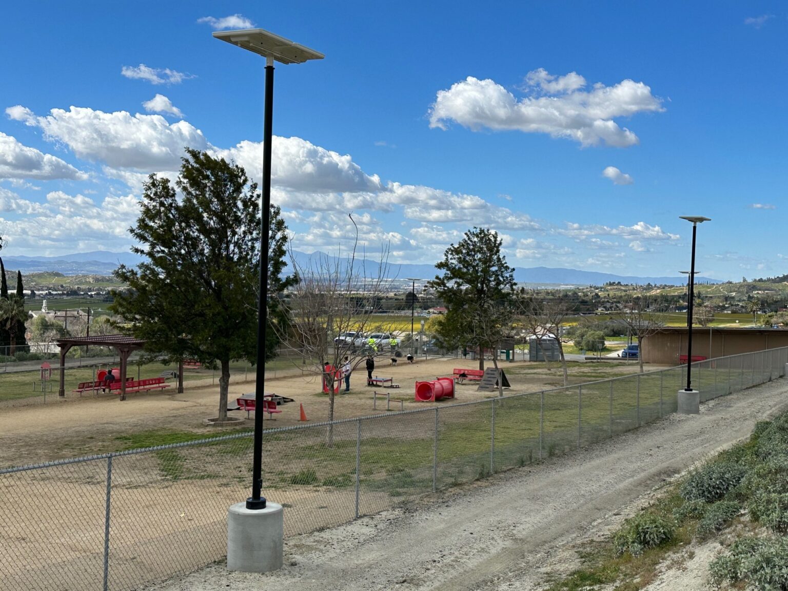 Two of Sol's iSSL solar lights at a dog park with agility equipment, trees, and covered areas with benches