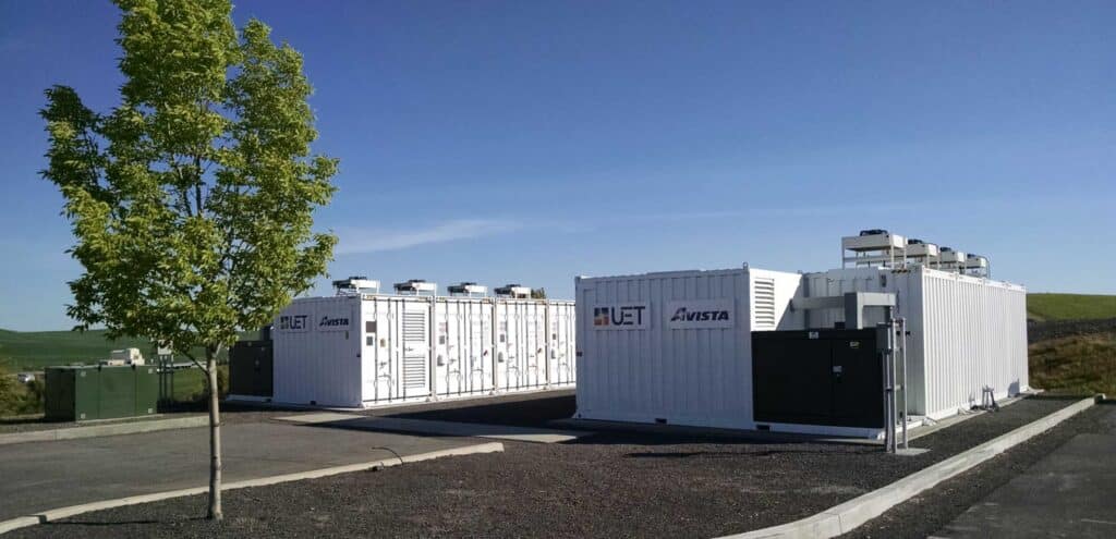 two-part battery energy storage system surrounded by asphalt and grass and a tree in the foreground